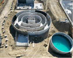 An aerial view of a large, circular oxidation ditch under construction. The ditch has multiple concrete walls and a central section with a water channel. There is also a smaller, circular tank filled with blue water nearby.