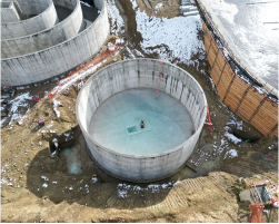 Aerial view of a large, circular concrete water tank under construction. The tank is surrounded by dirt and excavation equipment. Other smaller tanks can be seen in the background.